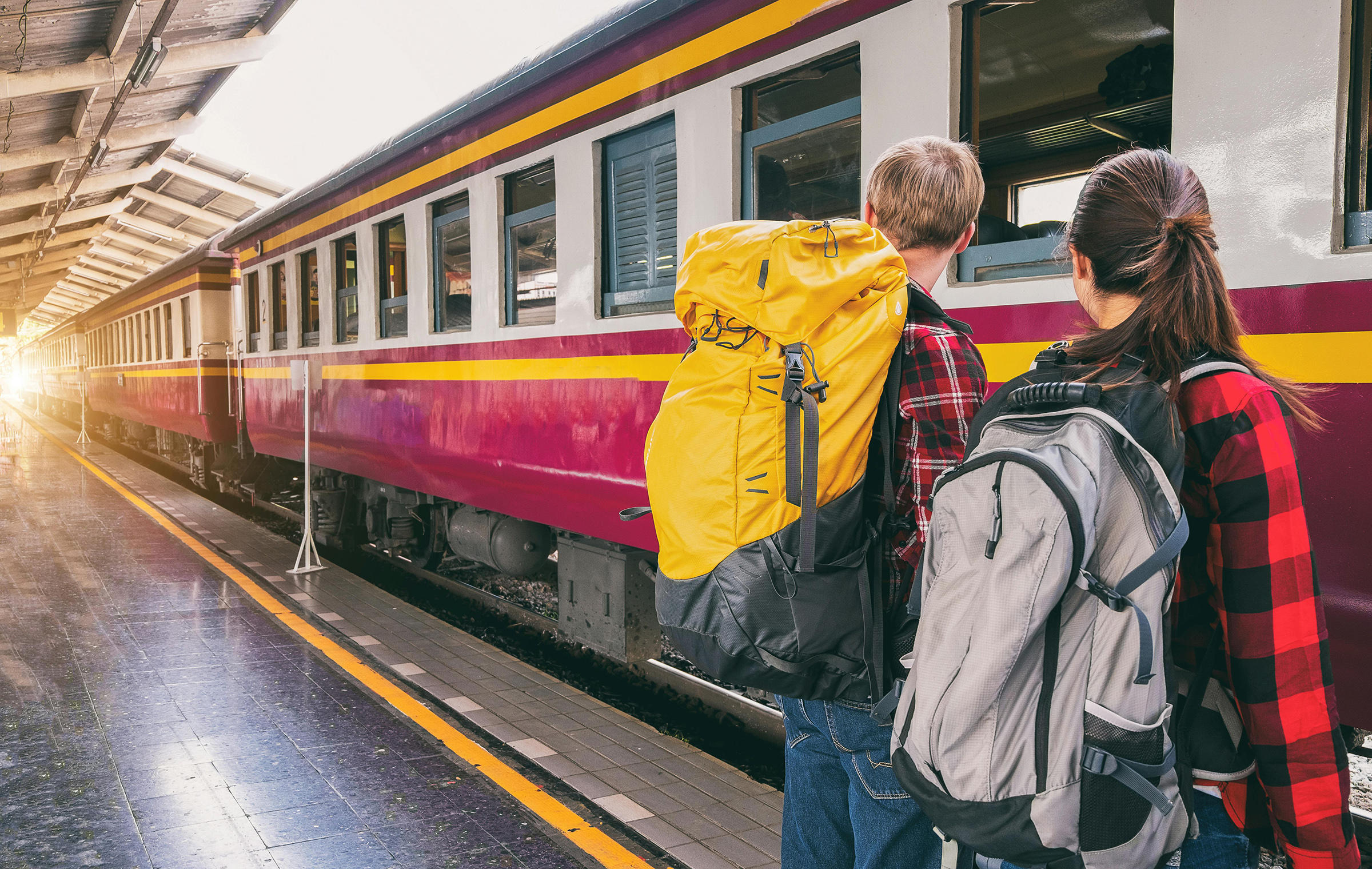 2 Jugendliche mit Rucksack von hinten fotografiert, die vor einem Zug am Bahnhof stehen.