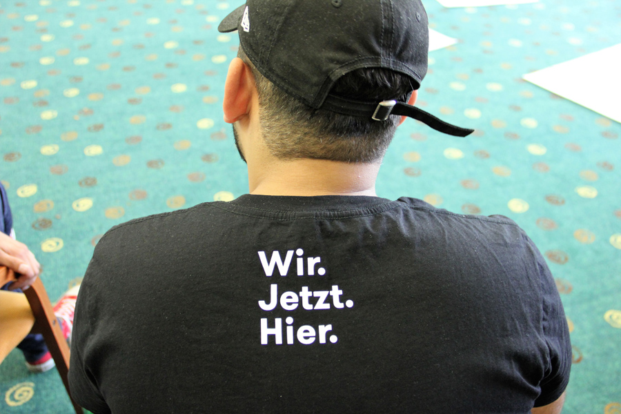 Rücken und Hinterkopf eines Mannes mit schwarzem T-Shirt, auf dem steht "Wir. Jetzt. Hier."