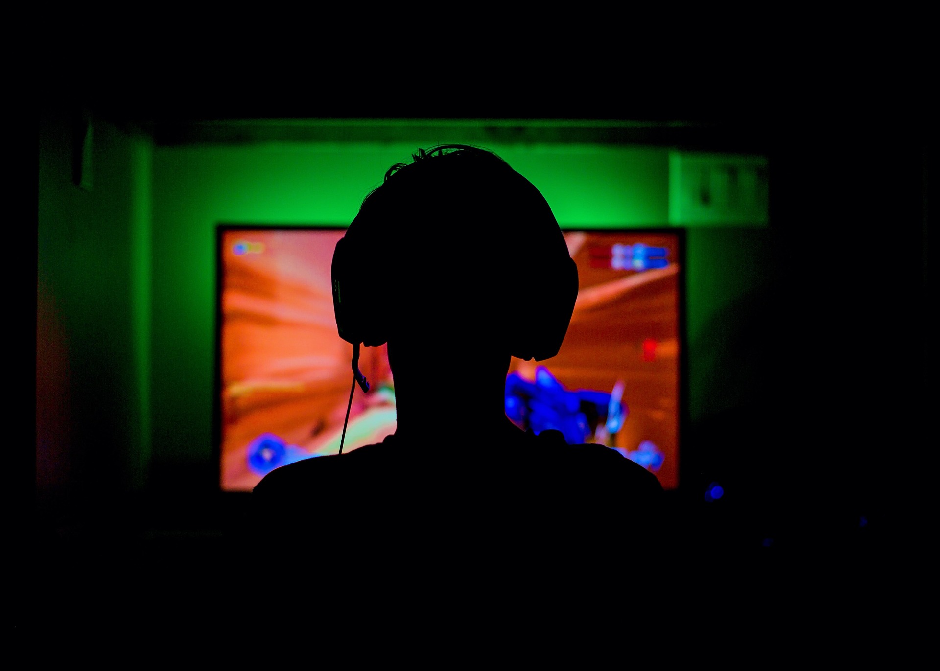 Silhouette eines jungen Mannes, der Kopfhörer auf hat und vor einem Screen sitzt und Computer spielt.