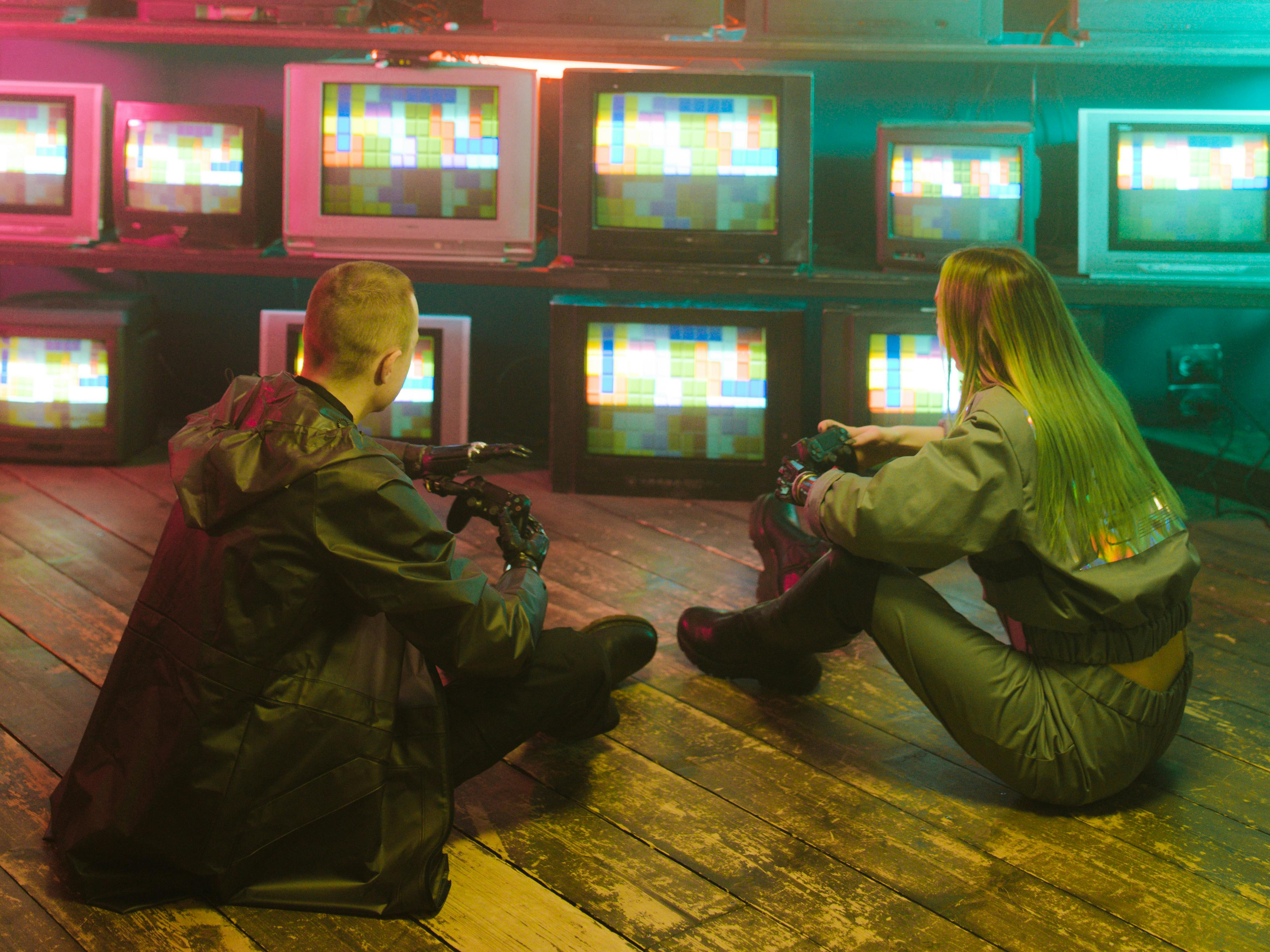 Zwei Jugendliche sitzen am Boden vor einer Wand mit vielen Monitoren.