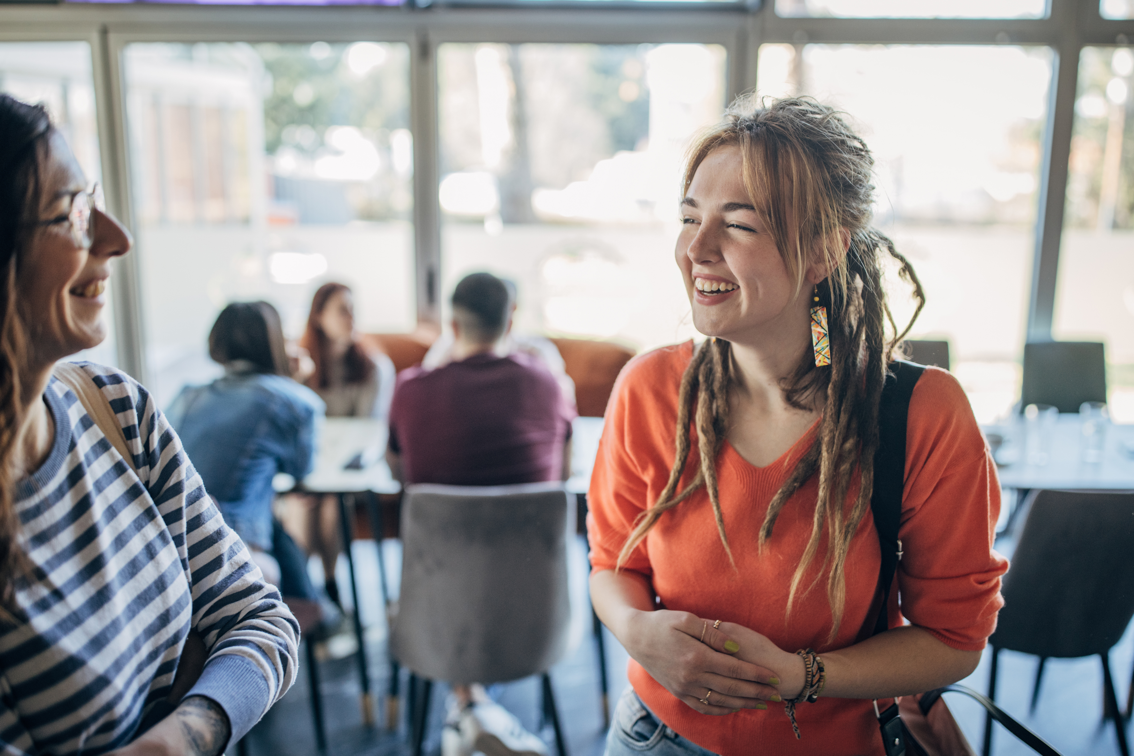 Junge Frau mit Dreadlocks und orangem Shirt lächelt und unterhält sich mit einer zweiten Frau in einem Cafe.