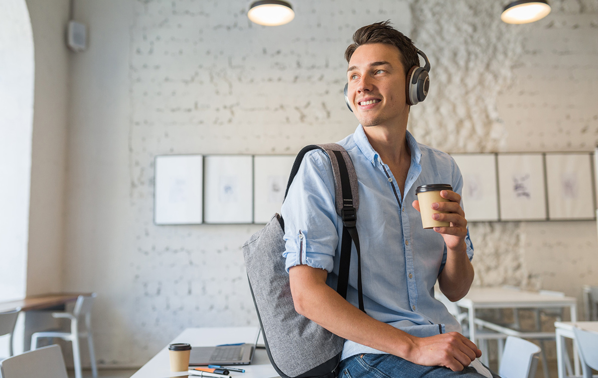 Junger Mann sitzt auf einem Tisch in einer Agentur, er hat Kopfhörer auf und einen Kaffeebecher in der Hand.