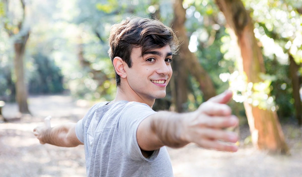 Junger Mann im Park, der gerade eine Dehnübung mit seinen Armen macht und in die Kamera lächelt.